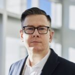 Profilporträt eines Geschäftsmannes in Anzug mit Brille in einem hellen Büro.